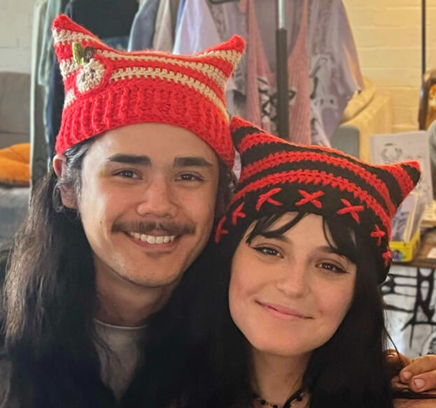 Image shows 2 people. One is wearing an apple themed crochet cat hat and the other is wearing a black and red 'x' crochet cat hat